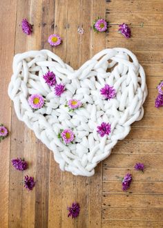 a white heart shaped basket with purple flowers in it on a wooden floor surrounded by petals