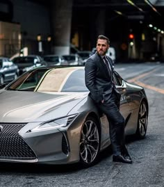 a man in a suit leaning on the hood of a silver sports car while standing next to it