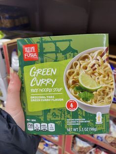 a person holding up a box of green curry ramen in front of some shelves