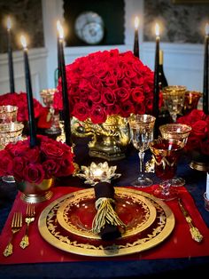 the table is set with red roses and gold place settings for two people to sit at