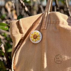 a sunflower pin sitting on the side of a tan tote bag that is hanging from a tree