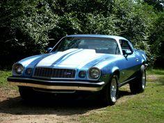 a blue and white car is parked on the side of a dirt road in front of some trees