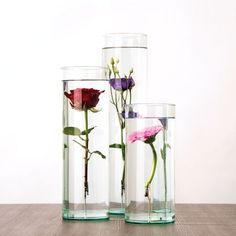 three glass vases with flowers in them on a wooden table next to a white wall