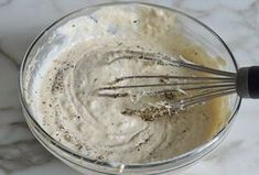 a glass bowl filled with white sauce and whisk on top of a marble counter