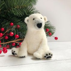 a small white polar bear sitting next to a christmas tree with red berries on it