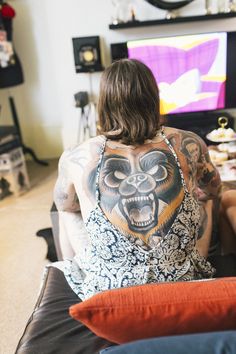 a man with tattoos on his back sitting in front of a flat screen tv and watching television