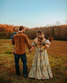 a man and woman holding hands while walking through a field with a baby in her arms