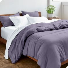 a bed with purple sheets and pillows in a white room next to a potted plant