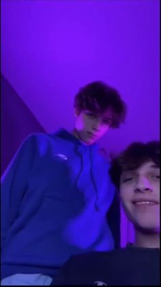 two young men standing next to each other in front of a purple wall and one is wearing a blue hoodie