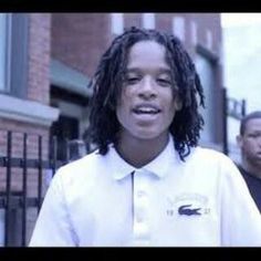 a young man with dreadlocks standing in front of a gate and another person behind him