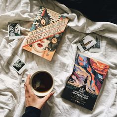 a person is holding a cup of coffee and reading books on a bed with white sheets