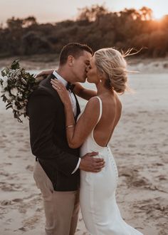 a bride and groom kissing on the beach at sunset with trees in the back ground