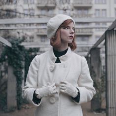 a woman with red hair wearing a white coat and black turtle neck sweater standing in front of a building