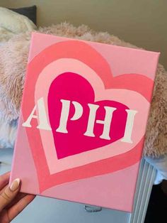 a person holding up a pink heart with the word phi painted on it in front of a teddy bear