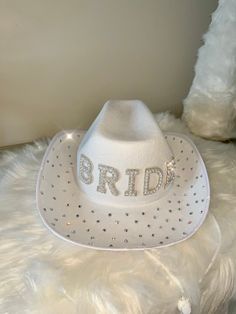 Grab this Adorable White Cowboy style Hat! Featuring BRIDE in silver and pearl decorations with the rim of the hat covered in scattered crystals. This Western style novelty hat is so fun! It is perfect for Night or a Bachelorette Party! Sized approximately 62 cm, it's great for both Men and Women! Wear it on your Wedding Day! Stylish and bedazzling, this Country Style hat is great for Rodeo style weddings, bridal shower supplies, and more!  The cute hat is lightweight and made of soft polyester. Spot clean only. White Bride Cowboy Hat, White Bedazzled Cowboy Hat, White Hats With Rhinestones And Curved Brim, White Rhinestone Hat With Curved Brim, Silver Wide Brim Costume Hat With Adjustable Fit, Silver Wide Brim Adjustable Costume Hat, Silver Adjustable Wide Brim Costume Hat, Silver Hat With Rhinestones For Rodeo, Western Silver Hat With Rhinestones