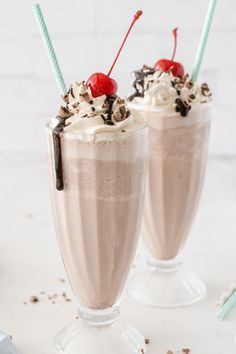 two glasses filled with chocolate milkshake and topped with whipped cream and cherries