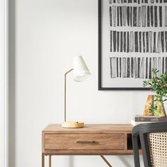 a wooden desk with a lamp on it next to a chair and framed art above it