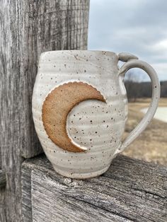 a ceramic mug with a half moon on it sitting next to a wooden fence post