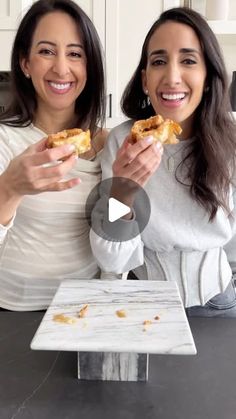 two women sitting at a kitchen counter eating food from their hands and posing for the camera