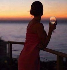 a woman in a red dress holding a glass of wine at the sunset overlooking the ocean