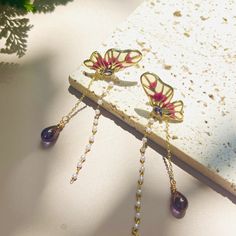 These exquisite butterfly earrings 🦋 feature delicate, golden butterflies with vibrant red and yellow wings, highlighted by a central gemstone 💎. Dangling from each butterfly is a chain of small, white beads, adding a touch of elegance 🌟. At the end of the chain, a stunning purple teardrop gemstone completes the look. These earrings are perfect for adding a whimsical and elegant flair to any outfit, making them a unique and charming addition to any jewelry collection 🌸. Handcrafted with meti Fairycore Butterfly Earrings For Gift, Gold Fairy Earrings For Gift, Red Butterfly Earrings For Gift, Fairycore Gold Drop Earrings, Gold Fairycore Drop Earrings, Gold Fantasy Earrings As Gift, Gold Fantasy Earrings For Gift, Fantasy Gold Earrings For Gift, Burgundy Butterfly