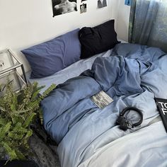 an unmade bed with blue sheets and black headphones on the bottom, next to a plant