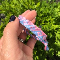 a hand holding a pink, blue and white braided bracelet in front of some bushes