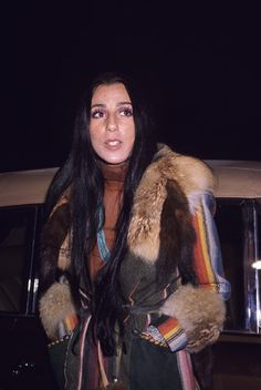 a woman standing in front of a car wearing a fur coat and holding a drink