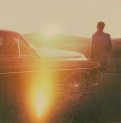 a man standing next to a parked car in the grass with the sun behind him