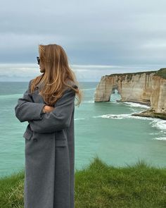 a woman standing on top of a lush green hillside next to the ocean with an arch in the background