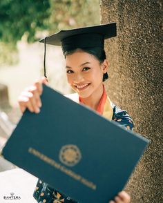 a woman wearing a graduation cap and gown holding up a blue book in front of a tree