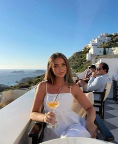 a woman holding a wine glass sitting on top of a table next to the ocean