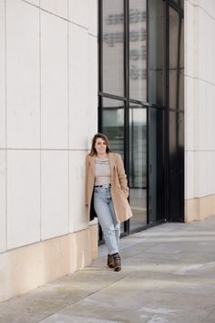 a woman leaning against the side of a building with her hand in her pocket and looking at the camera