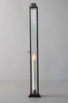 a candle is lit in a tall glass container on a table with a white wall behind it