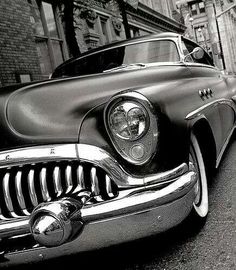 black and white photograph of an old car parked on the side of the road in front of a brick building