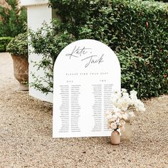 a white sign sitting on top of a lush green field next to a flower filled planter