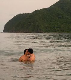 two people are kissing in the water near some hills and an island with green trees