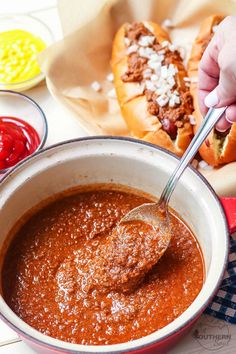 a person spooning hot dog sauce into a pot with chili and cheese on the side