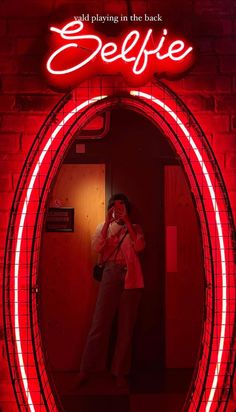 a woman taking a selfie in front of a red neon sign that says selfie