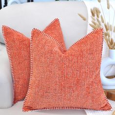 an orange pillow sitting on top of a white couch next to a vase with dry grass