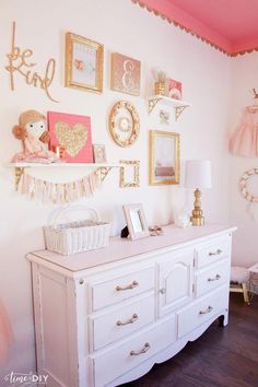 a white dresser topped with lots of pictures next to a wall filled with pink and gold decorations