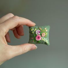 a hand is holding a small embroidered flowered pillow in front of a gray background