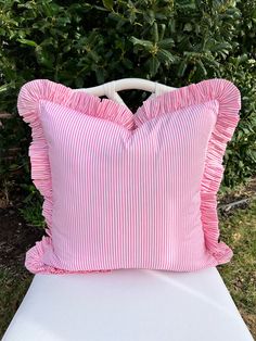 a pink and white striped pillow sitting on top of a white chair in front of some bushes