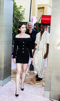 two men and a woman are standing in front of an open door, one is wearing a black off the shoulder dress