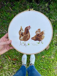 a person holding up a cross - stitch with two chickens on it's side