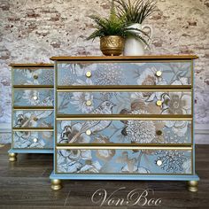 a blue and gold dresser with flowers painted on the drawers, next to a potted plant