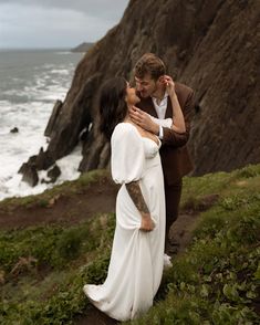 a man and woman standing next to each other in front of the ocean on a cliff