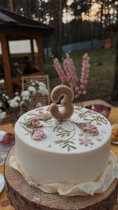 a white cake sitting on top of a tree stump covered in flowers and the number three