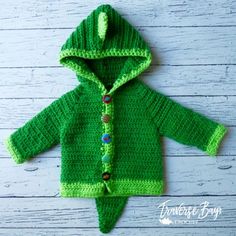 a green crocheted jacket with hood and buttons on the front, sitting on a white wooden surface