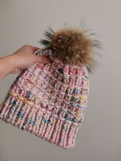 a hand holding a knitted hat with a fur pom